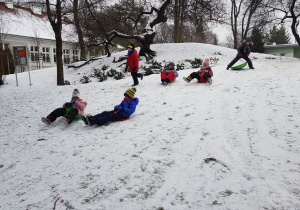 Grupa dzieci ślizga się na ośnieżonej górce w ogrodzie przedszkolnym